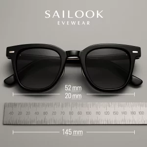 A close-up of Sailook Eyewear sunglasses featuring sleek black acetate frames with gold-tone accents, dark grey gradient lenses, and a 52 mm frame width. The temple length of 145 mm is marked on a metal ruler below the sunglasses, set against a soft grey background.