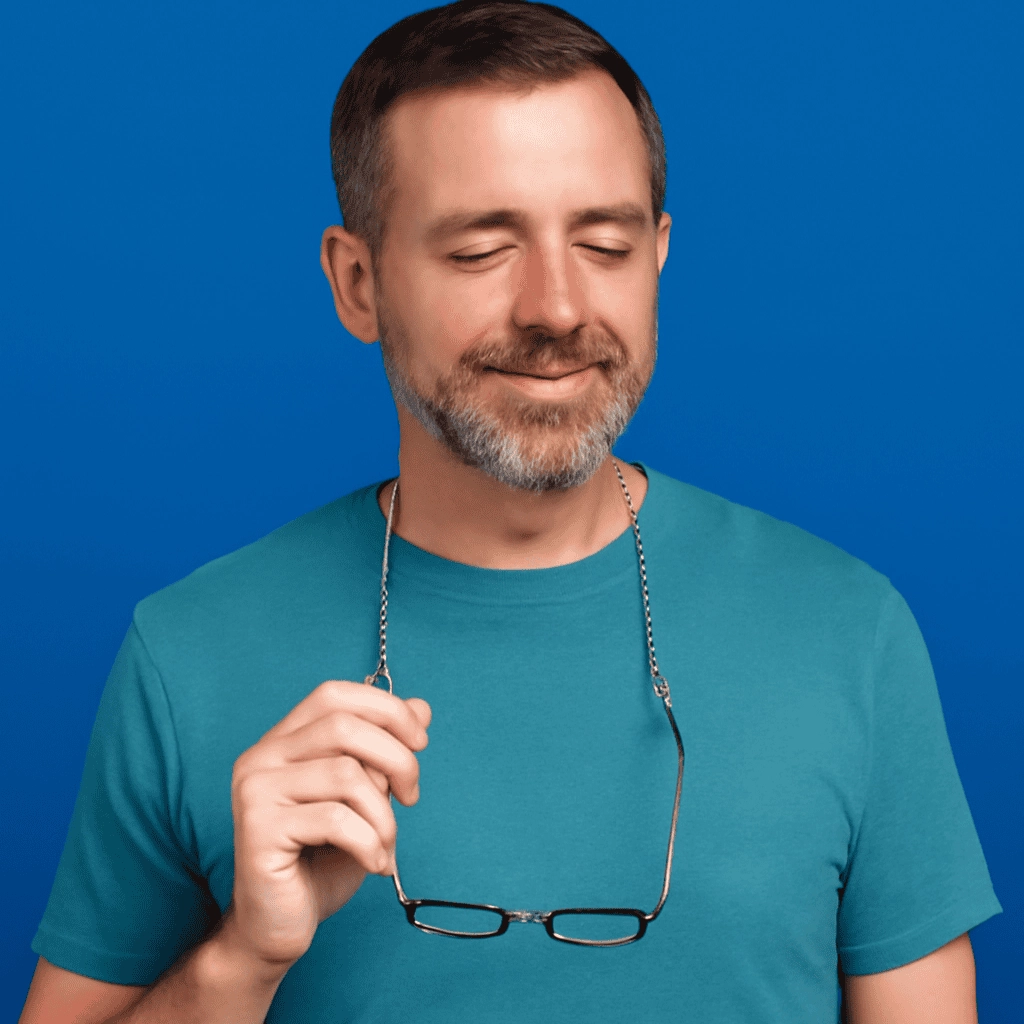 A middle-aged man with short brown and gray hair, a well-groomed beard, and light skin, smiling gently with eyes closed, holding reading glasses around his neck on a silver chain. He wears a teal T-shirt, with a soft blue background.