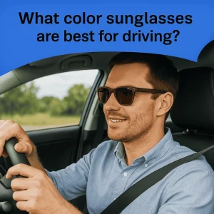 A man in his 30s, wearing brown polarized sunglasses, smiles while driving a car. He is dressed in a light blue shirt with a relaxed grip on the steering wheel. The background shows a blurred green landscape under a clear blue sky, with the text 'What color sunglasses are best for driving?' in bold above.