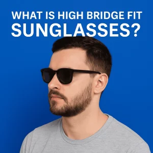 A bearded man with light skin and short dark hair wears high bridge fit sunglasses against a solid blue background. White bold text above him reads, "WHAT IS HIGH BRIDGE FIT SUNGLASSES?" His sunglasses sit high and evenly on his nose bridge, illustrating the concept visually.