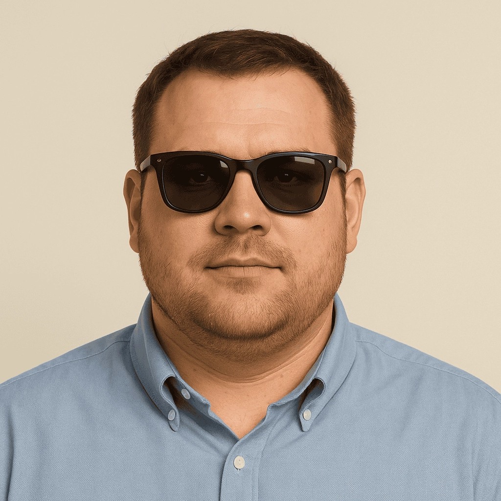 A head-and-shoulders portrait of a light-skinned man with a broad face wearing black wayfarer sunglasses and a light blue button-up shirt against a neutral beige background.